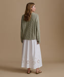 Woman wearing a green cable knit sweater and white dress with cut-out patterns on a beige background