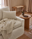 A cozy white sofa with the Alpaca Basketweave Lumbar Pillow and a knitted blanket sits beside a round wooden side table displaying a dark vase with flowers and a decorative dish, while a wooden bench and beige curtains enhance the inviting room.