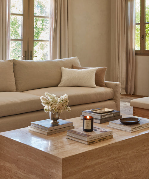A cozy living room with a beige sofa, the Alpaca Basketweave Lumbar Pillow, and a throw blanket. A travertine coffee table holds stacked books, a candle, a small vase of white flowers, and a ceramic dish. Large windows fill the space with natural light.