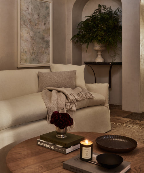 A cozy living room with a cream sofa, the Alpaca Basketweave Lumbar Pillow, and a beige throw. A wooden coffee table holds books, a candle, dark flowers, and ceramics. Artwork, a plant, and an arched alcove accent the softly lit space.