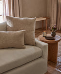 A cozy beige sofa with an Alpaca Basketweave Lumbar Pillow sits beside a wooden side table holding a lit candle, a small vase of flowers, and a dark dish in a softly lit room with stone flooring and neutral curtains.