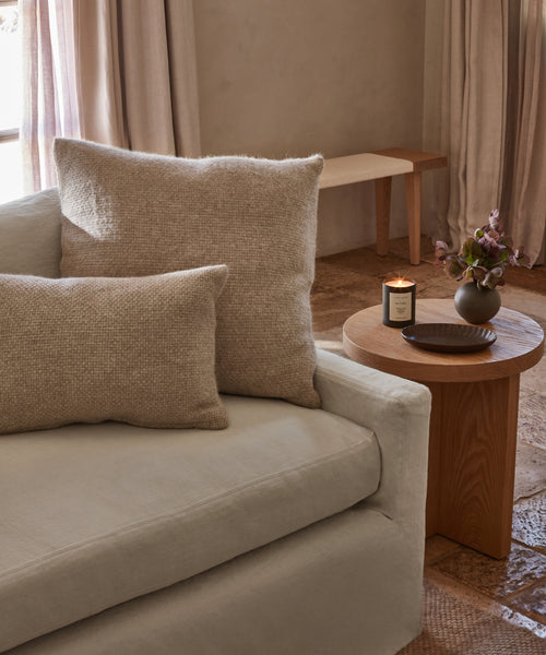 A cozy beige sofa with an Alpaca Basketweave Lumbar Pillow sits beside a wooden side table holding a lit candle, a small vase of flowers, and a dark dish in a softly lit room with stone flooring and neutral curtains.