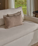 A cream-colored sofa is paired with two beige pillows, including the Alpaca Basketweave Lumbar Pillow—handwoven in Peru—by a window with white curtains and natural light. A small round wooden side table sits next to the sofa.