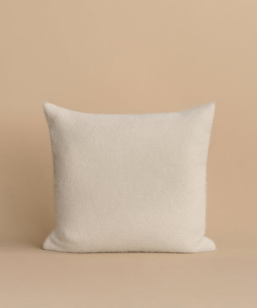 The Alpaca Basketweave Pillow, a square white accent, is handmade in Peru and sits upright on a beige surface with a matching beige background for an elegant touch.