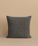 The Alpaca Basketweave Pillow, square-shaped with a textured dark gray design, is displayed upright against a plain light beige background.