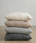 A stack of four neutral-tone pillows, featuring the Alpaca Basketweave Pillow for added softness, rests on a light wooden floor against a plain white wall, blending modern simplicity with cozy luxury.