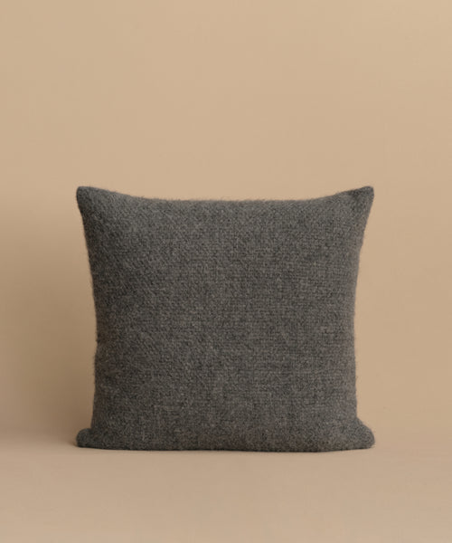 The Alpaca Basketweave Pillow, square-shaped with a textured dark gray design, is displayed upright against a plain light beige background.