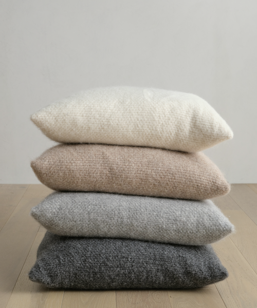 A stack of four neutral-tone pillows, featuring the Alpaca Basketweave Pillow for added softness, rests on a light wooden floor against a plain white wall, blending modern simplicity with cozy luxury.