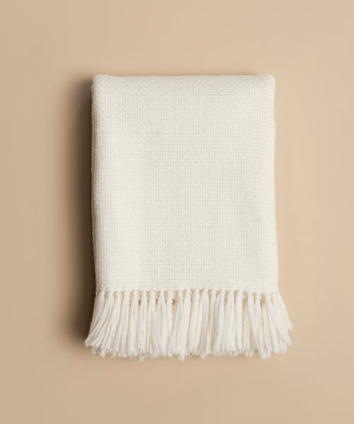 The Alpaca Basketweave Throw, handwoven in Peru, features an off-white color with fringe and is neatly folded on a neutral beige background.