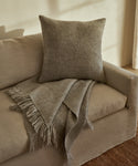 A beige sofa with a textured gray cushion and a Large Alpaca Basketweave Throw, handwoven in Peru, draped over the armrest in a softly lit room with neutral tones.