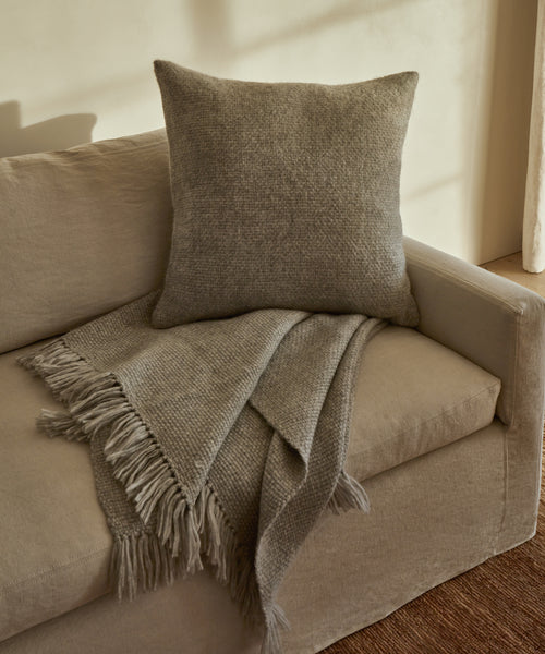 A beige sofa with a textured gray cushion and a Large Alpaca Basketweave Throw, handwoven in Peru, draped over the armrest in a softly lit room with neutral tones.