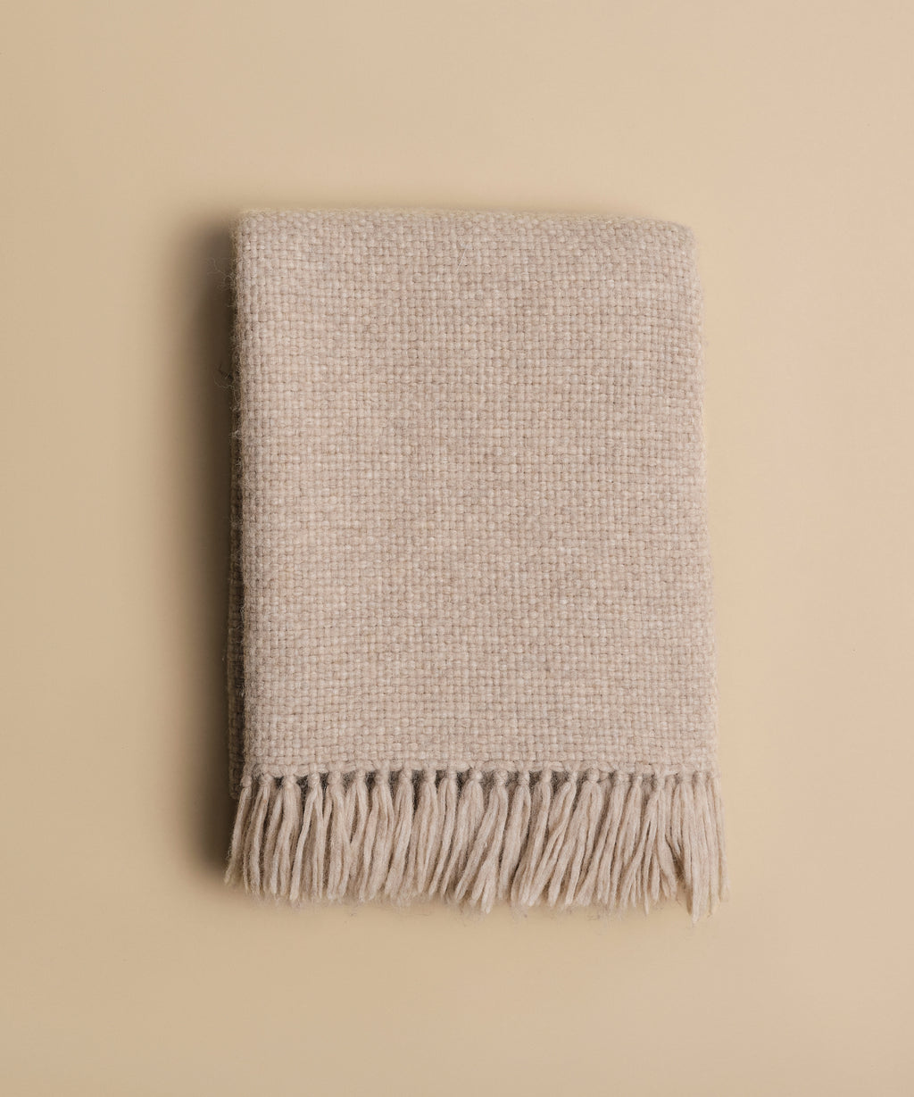 The Alpaca Basketweave Throw, in beige with fringed edges, is folded on a plain, light tan background, highlighting its super-soft, handwoven Peruvian texture.