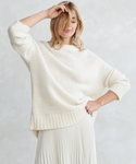A woman models the Alpaca Cocoon Crewneck in cream, paired with a pleated white skirt, posing against a light textured background with one hand on her hip and the other partly covering her face.