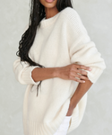 A woman stands against a light gray background, arms crossed, wearing the Alpaca Cocoon Crewneck—a cozy, oversized chunky sweater with long sleeves. Her long, braided hair drapes over her shoulder.