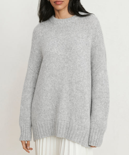 A woman stands against a plain background wearing the Alpaca Cocoon Crewneck—a loose-fitting, light gray sweater with long sleeves and a ribbed hem—paired with a white pleated skirt.