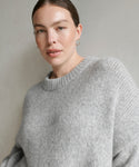 A person with light skin and brown hair pulled back wears the Alpaca Cocoon Crewneck sweater in grey with small hoop earrings, gazing confidently at the camera against a light background.