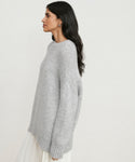 A woman with long dark hair wears the Alpaca Cocoon Crewneck, a loose-fitting light gray sweater, paired with a white pleated skirt as she stands in profile against a plain background.