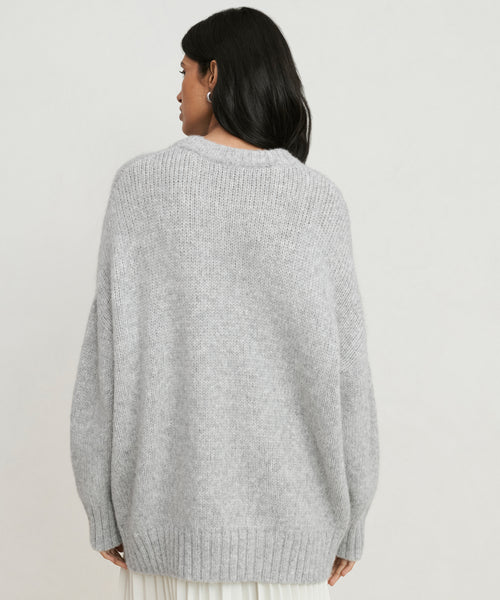 A woman with long dark hair is seen from the back, wearing the Alpaca Cocoon Crewneck—a loose light gray sweater with ribbed cuffs and hem—against a plain light background.