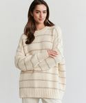A woman with long brown hair wears the Alpaca Cocoon Crewneck, an oversized sweater featuring subtle horizontal beige stripes, standing against a plain light background with her arms gently crossed.