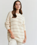 A woman with long brown hair models the Alpaca Cocoon Crewneck, a cream-colored striped sweater, paired with light pants as she poses confidently against a plain light background.
