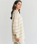 A woman with long brown hair stands sideways against a plain white background, wearing the Alpaca Cocoon Crewneck—a loose, cream sweater with light beige stripes—paired with cream pants.