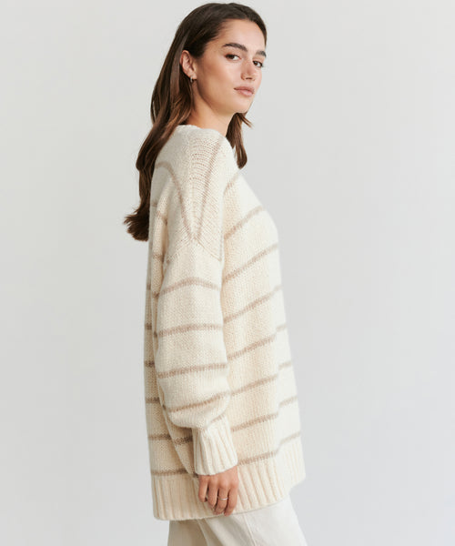 A woman with long brown hair stands sideways against a plain white background, wearing the Alpaca Cocoon Crewneck—a loose, cream sweater with light beige stripes—paired with cream pants.