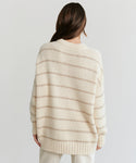 A person with long brown hair faces away, wearing the Alpaca Cocoon Crewneck—a loose, cream sweater with thin beige stripes and ribbed trim—paired with light pants against a plain background.