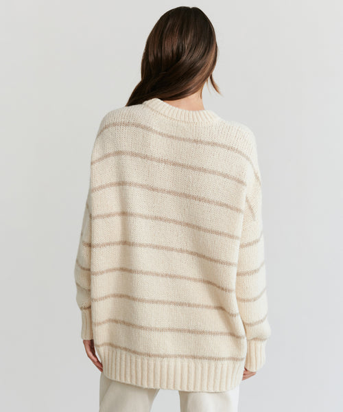 A person with long brown hair faces away, wearing the Alpaca Cocoon Crewneck—a loose, cream sweater with thin beige stripes and ribbed trim—paired with light pants against a plain background.