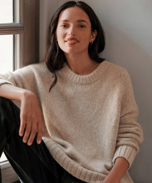 A woman with long dark hair, dressed in the Alpaca Cocoon Crewneck and black pants, sits by a window, smiling softly at the camera. Soft light enhances the cozy, inviting feel of her look.