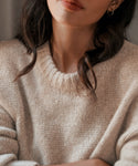 A woman with wavy brown hair wears the Alpaca Cocoon Crewneck, a light beige chunky sweater, paired with a small hoop earring; her face is partially out of view.