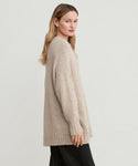 A woman with long, light brown hair models the Alpaca Cocoon Crewneck—a loose, chunky beige knit sweater—paired with black pants, standing sideways and looking over her shoulder against a plain white background.