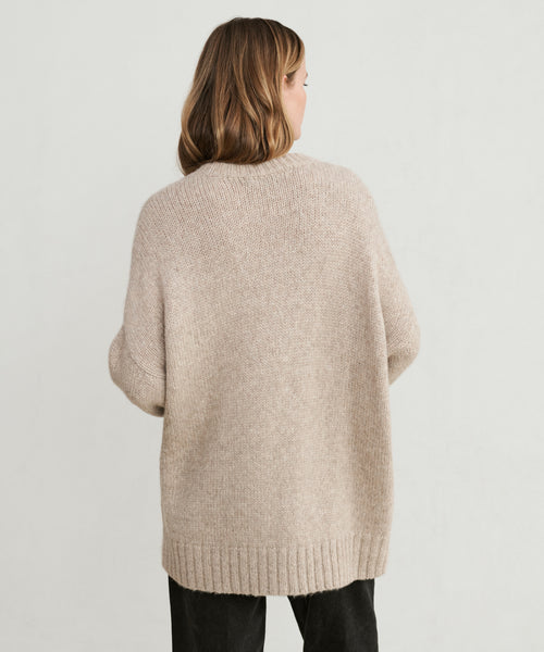 A woman with shoulder-length light brown hair is seen from behind, wearing the Alpaca Cocoon Crewneck in beige and black pants, standing against a plain light background.