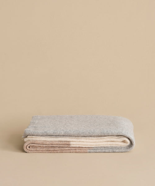 A neatly folded Alpaca Colorblock Throw in soft grey and beige rests on a neutral surface and matching background, highlighting the luxury of this handmade Peruvian piece.