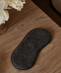 An Alpaca Eye Mask in dark gray rests on a wooden surface beside a vase of white flowers.