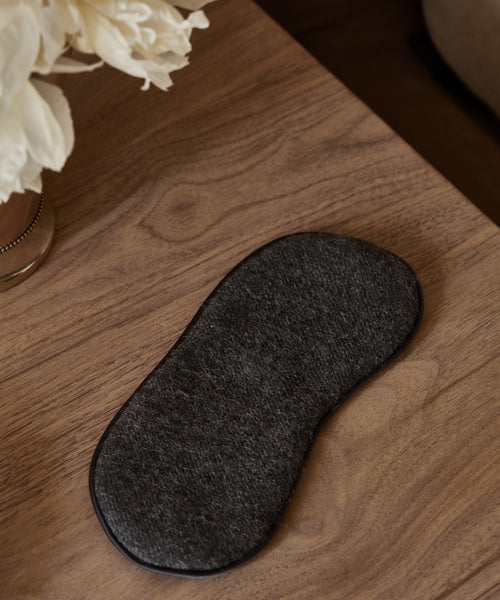 An Alpaca Eye Mask in dark gray rests on a wooden surface beside a vase of white flowers.