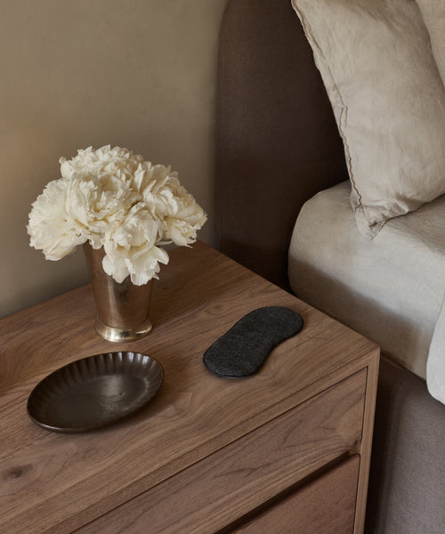 A wooden bedside table holds a metal vase of white flowers, a brown dish, and an Alpaca Eye Mask, beside a brown upholstered bed with beige bedding.