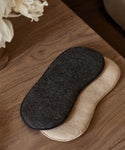 Two Alpaca Eye Masks, one in dark gray and one in beige, are stacked on a wooden surface near a bouquet of white flowers.