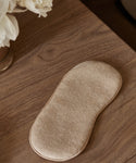 An Alpaca Eye Mask in beige alpaca wool rests on a wooden surface next to a vase of white flowers.
