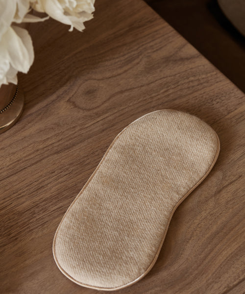 An Alpaca Eye Mask in beige alpaca wool rests on a wooden surface next to a vase of white flowers.