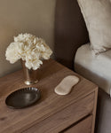 A wooden bedside table holds a beige Alpaca Eye Mask, a silver vase of white flowers, and a dark ceramic dish. Light bedding from the adjacent bed is also visible.
