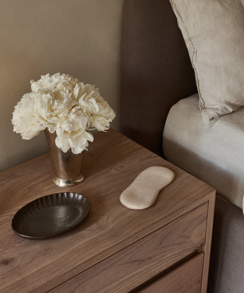 A wooden bedside table holds a beige Alpaca Eye Mask, a silver vase of white flowers, and a dark ceramic dish. Light bedding from the adjacent bed is also visible.
