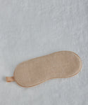 The Alpaca Eye Mask in beige, featuring a soft texture, is shown lying flat on a white fabric surface.
