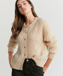 A woman with long brown hair wears the Cable Crewneck Cardigan, a relaxed-fit textured knit with buttons, paired with black jeans. She stands hands-in-pockets against a light background, highlighting its effortless style.