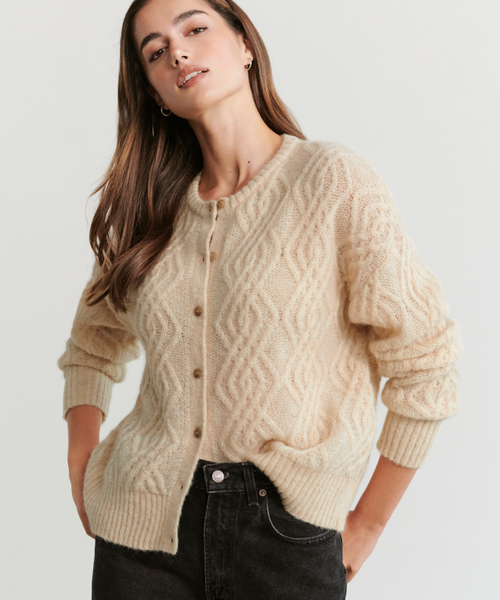 A woman with long brown hair wears the Cable Crewneck Cardigan, a relaxed-fit textured knit with buttons, paired with black jeans. She stands hands-in-pockets against a light background, highlighting its effortless style.