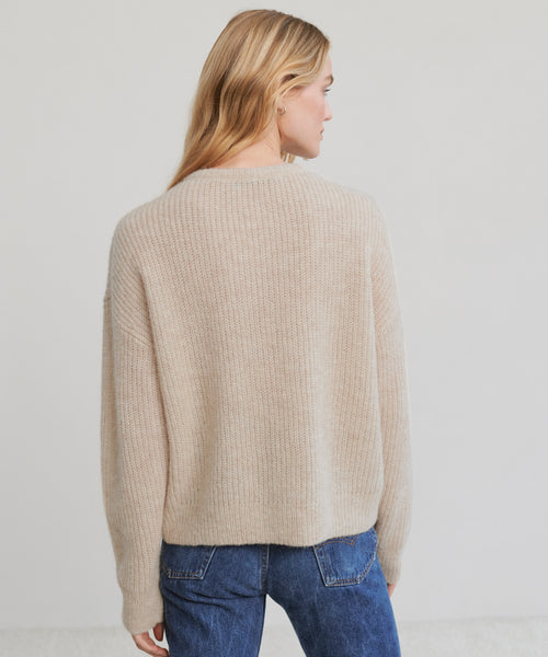 A woman with long blonde hair, seen from behind, wears the Hayes Crewneck Cardigan—a luxe fuzzy beige knit—paired with blue jeans against a plain light background.
