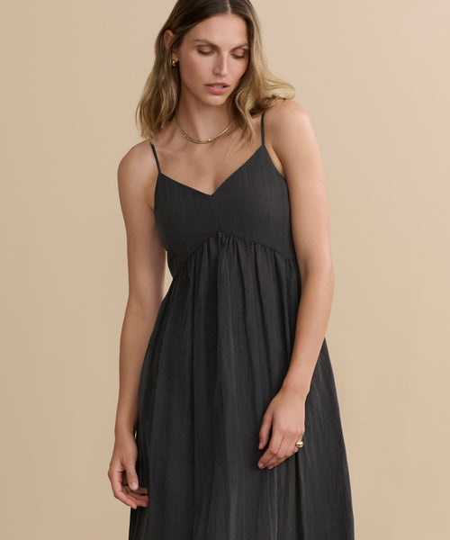 A woman wearing the Alyssandra Dress—a sleeveless, dark gray V-neck dress with thin straps and textured fabric—stands against a beige background, looking slightly downward with one arm by her side.