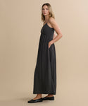 A blonde woman wears the Alyssandra Dress, a sleeveless dark gray maxi dress with textured fabric, paired with black flat shoes, standing sideways against a plain beige background.