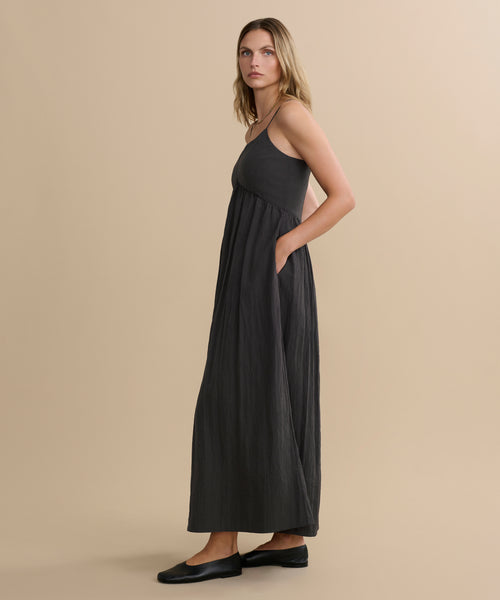 A blonde woman wears the Alyssandra Dress, a sleeveless dark gray maxi dress with textured fabric, paired with black flat shoes, standing sideways against a plain beige background.