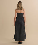 A woman with shoulder-length blonde hair stands facing away, wearing the Alyssandra Dress—a long, sleeveless black dress with thin straps—paired with black shoes, set against a plain beige backdrop.