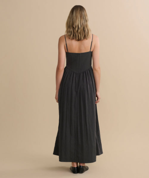 A woman with shoulder-length blonde hair stands facing away, wearing the Alyssandra Dress—a long, sleeveless black dress with thin straps—paired with black shoes, set against a plain beige backdrop.